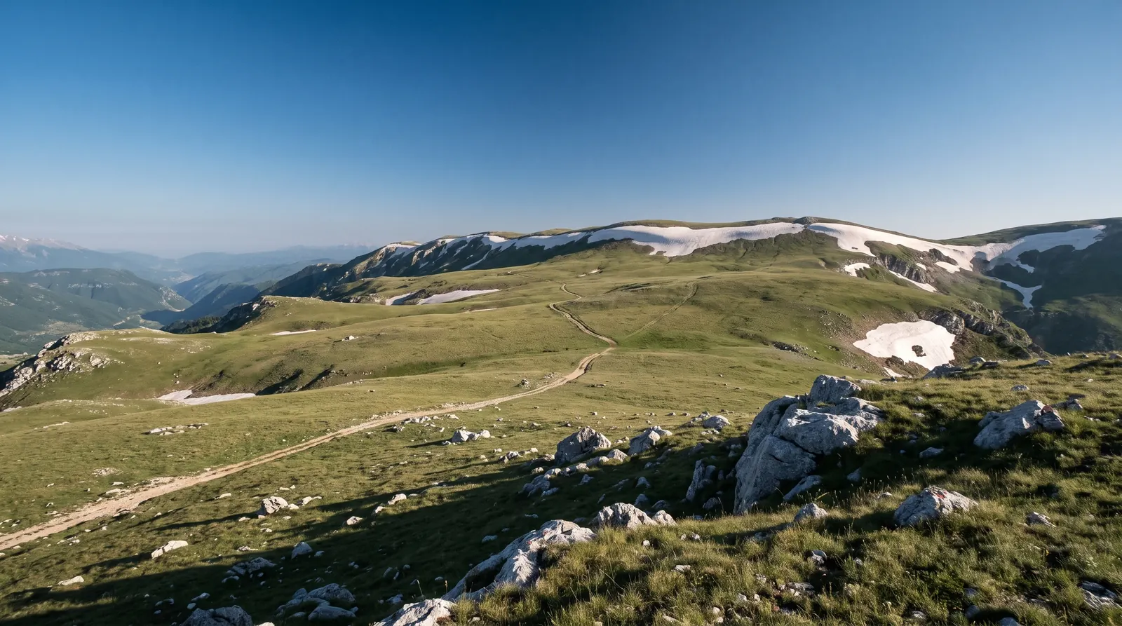 A mule path crossing the high meadows of Gramoz toward the summit ridge, late snow banks under the crest