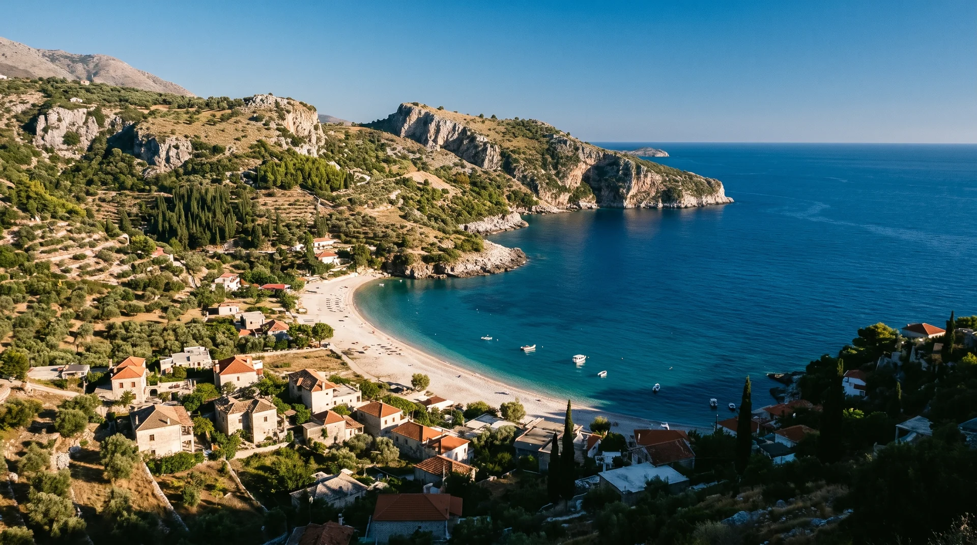 The curve of Himarë bay from an elevated viewpoint, pebble beach below olive and cypress hills